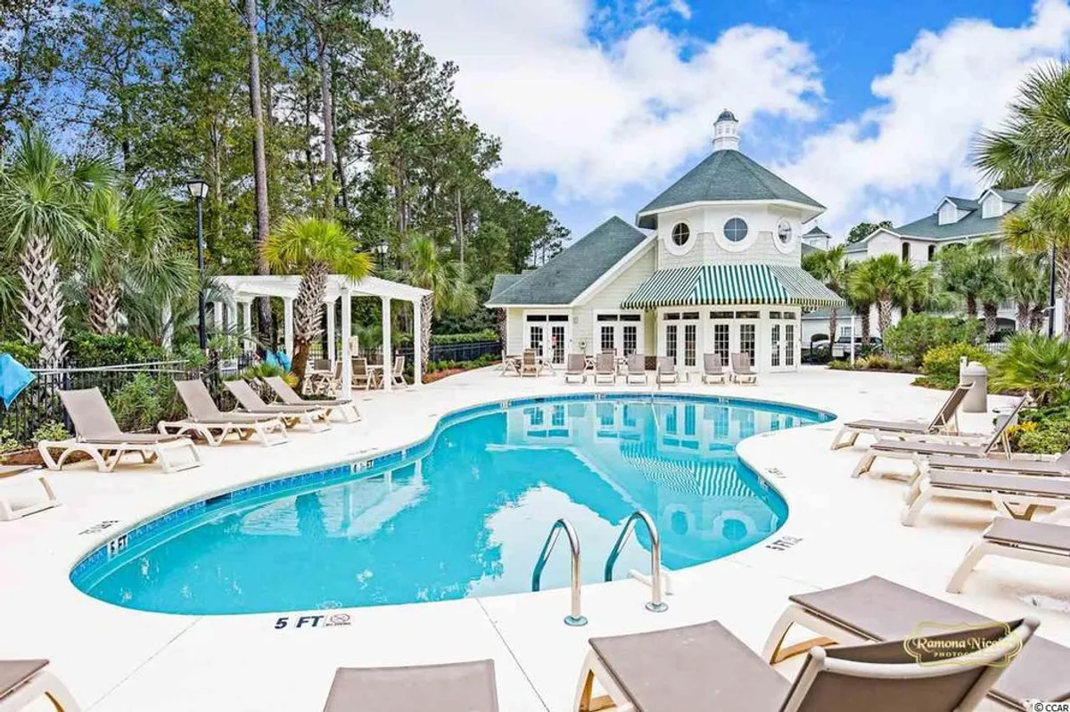 This image features the inviting swimming pool area surrounded by lounge chairs, landscaped greenery, and a view of the clubhouse. The scene conveys a leisurely resort atmosphere, ideal for both relaxation and social gatherings.