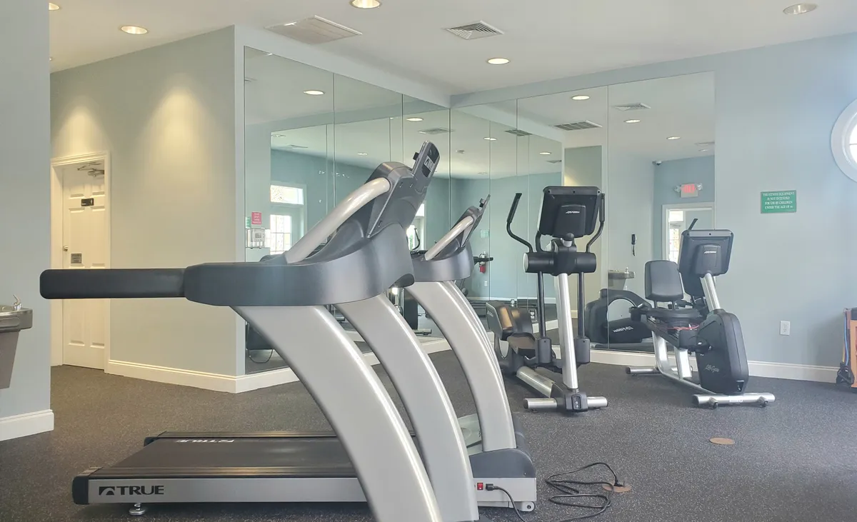 A bright and modern fitness center featuring a treadmill, elliptical machine, and exercise bike. The space is designed for both individual workouts and group fitness, with large windows that allow plenty of natural light.