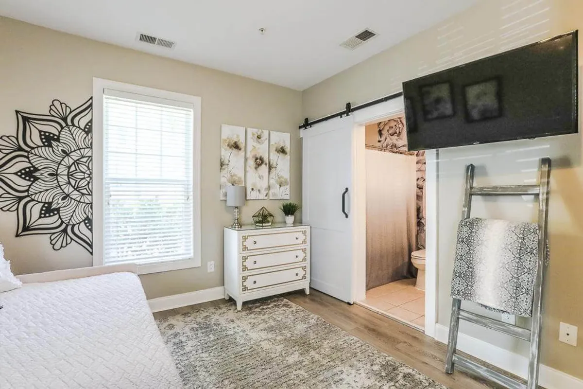 A serene bedroom with crisp white bedding, black mandala wall art, a white dresser, decorative plants, and a flat-screen TV. The setting feels peaceful and perfect for intimate relaxation.