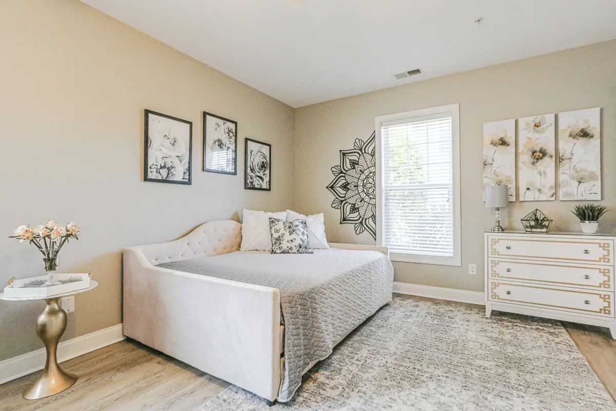 A cozy bedroom corner with a bed layered in soft linens and decorative pillows, framed floral artwork above a decorative dresser, and gentle lighting that creates a comfortable, serene environment perfect for relaxation or quiet reflection.