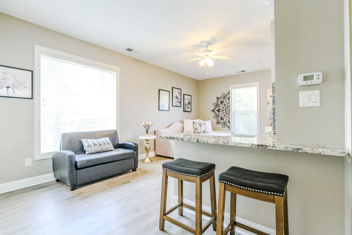 A bright, comfortable living space is shown, featuring a plush sofa, bar stools at the counter, vibrant wall art, and sunlit windows. The overall atmosphere is welcoming, ideal for moments of relaxation, reading, or enjoying light meals.