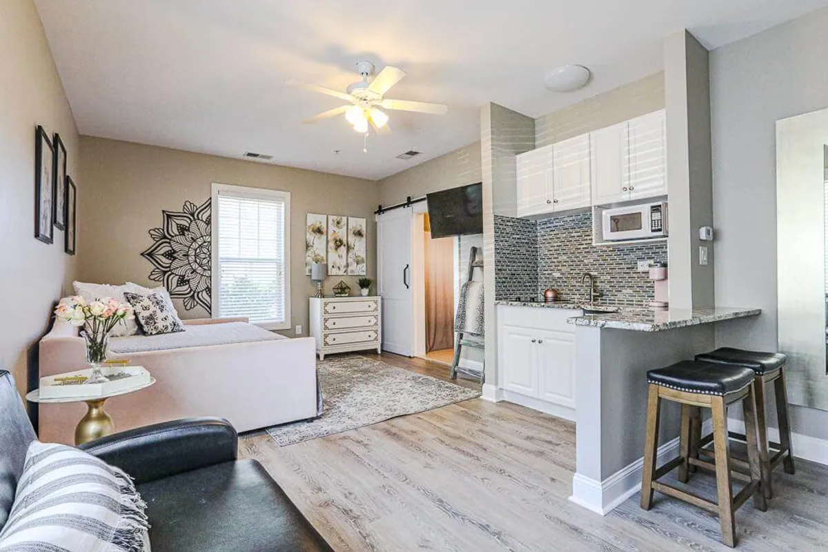 A welcoming living area that showcases a sofa with decorative pillows, a well-equipped kitchenette, a compact dining table with fresh flowers, and vibrant wall art. The scene suggests a multi-use space ideal for relaxing, socializing, or enjoying a meal.