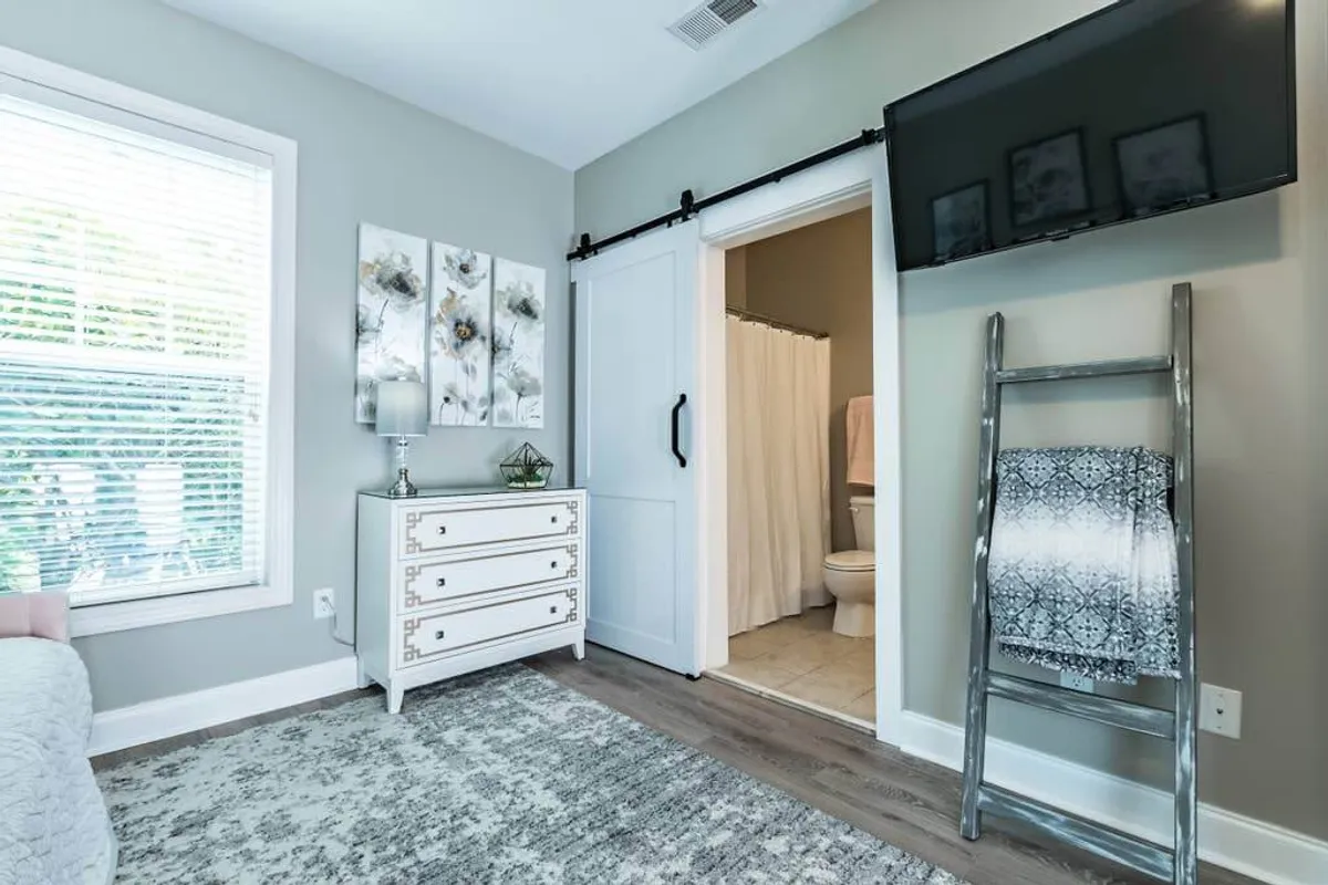 A serene studio bedroom with a neatly made bed featuring decorative pillows, a white dresser topped with floral artwork, a rustic sliding barn door, and a wall-mounted television. The space is bathed in soft natural light, enhancing the inviting atmosphere.