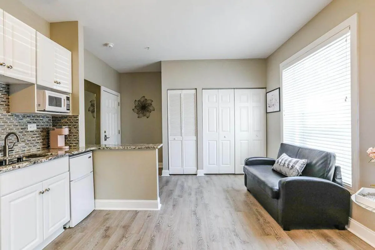 This image captures a cozy open-plan living area featuring a well-lit kitchen space, a comfortable sofa, a large window with blinds, and accessible closet doors. The scene hints at a welcoming environment where guests can relax or start their day with breakfast by the window.